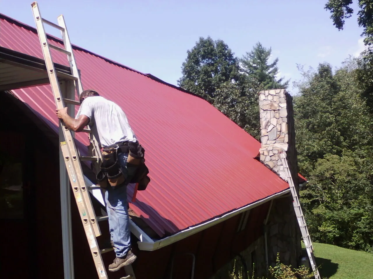 Roofer on a ladder beside a red metal roof and stone chimney — Soffit & Fascia Repair by Roofing Brothers in Falls